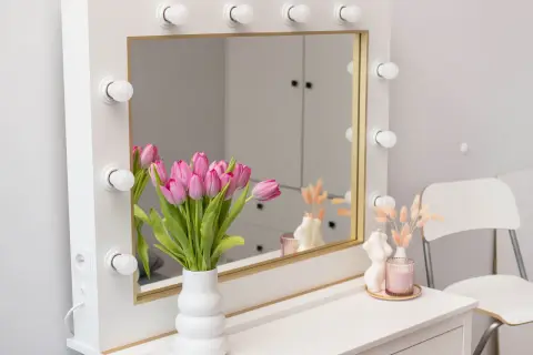 Stylowe lustro z lampkami, idealne do makijażu. Obok bukiet różowych tulipanów w wazonie.