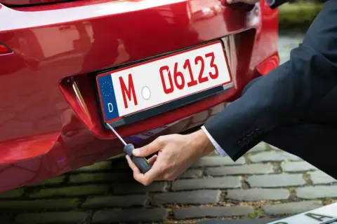 Mann schraubt Kennzeichen an rotes Auto. So bekommt man ein rotes Kennzeichen für Überführungsfahrten.