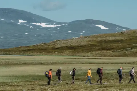Vandring i fjällandskap med snöklädda toppar, en grupp vandrare med ryggsäckar rör sig över en gräsbevuxen slätt mot Helags Sylarna.