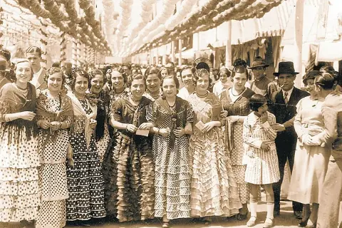 Grupo de mujeres y niñas con trajes de volantes y lunares, evocando el origen del traje de flamenca en una feria antigua.