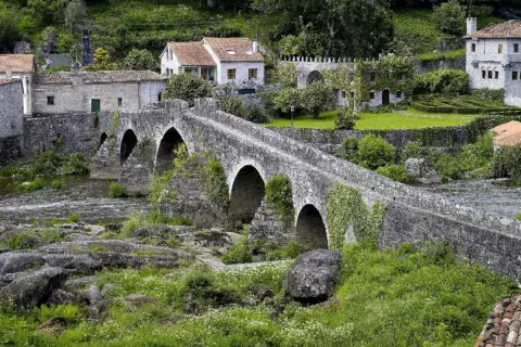Un antiguo ponte carreira de piedra cruza un río, rodeado de casas rústicas y vegetación exuberante.
