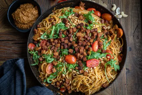 En stor panna med spaghetti blandad med krämig pesto rosso, toppad med krispiga tempebitar, pinjenötter, körsbärstomater och ruccola. Ett perfekt pesto rosso recept!