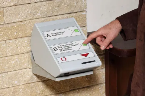 Dłoń naciska przycisk na automacie do wydawania dowodów rejestracyjnych, ułatwiając wymianę dowodu rejestracyjnego online.