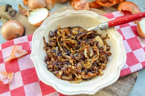 Eine Schüssel mit köstlichen, karamellisierten Zwiebeln, umgeben von frischen Zwiebeln und ihrer Schale. Perfekt als Beilage oder Topping.