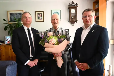 Trzech mężczyzn, jeden z bukietem kwiatów, w eleganckich strojach.
