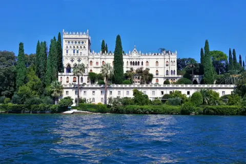 Villa neogotica su un'isola del Lago di Garda, circondata da cipressi e vegetazione lussureggiante, con il blu intenso dell'acqua in primo piano.