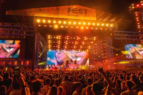 Tłum na festiwalu Fest, rozświetlony sceną i ekranami z artystami. Widać logo "FEST festival" i tłum ludzi z telefonami.
