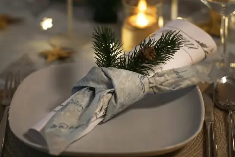Une serviette en tissu, décorée d'une branche de sapin et d'une pomme de pin, est pliée en forme de smoking sur une assiette.