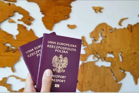 Dłoń trzyma dwa polskie paszporty na tle drewnianej mapy Europy. Czas na podróż!