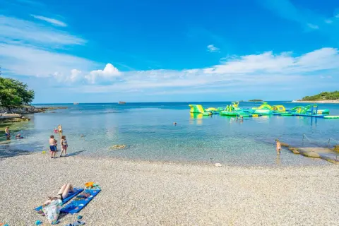 Słoneczna plaża z kamykami, ludzie kąpią się w krystalicznie czystej wodzie, a w oddali wodny plac zabaw. Idealne miejsce na wakacje w camping Bijela Uvala.