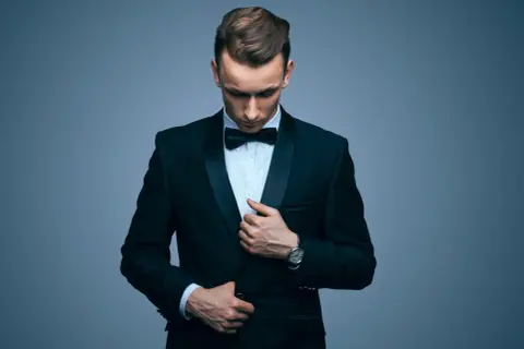 Elegancki mężczyzna w galowym stroju męskim, z czarną muszką i zegarkiem na ręku, poprawia marynarkę.