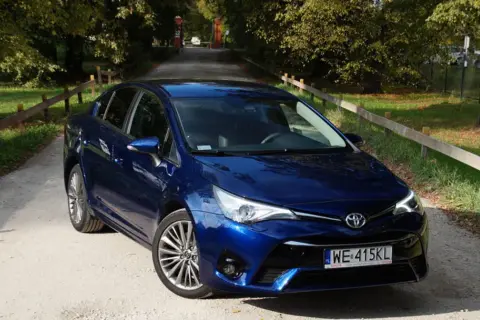 Błękitna Toyota Avensis T27 na żwirowej drodze, otoczona zielenią i drzewami.