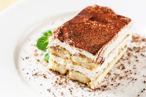 Une part de tiramisu mascarpone, aérienne et gourmande, saupoudrée de cacao, avec une touche de menthe fraîche.