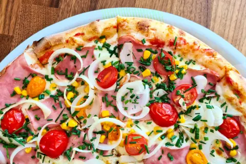 Zbliżenie na pizzę z szynką, pomidorkami koktajlowymi, kukurydzą, cebulą i szczypiorkiem.