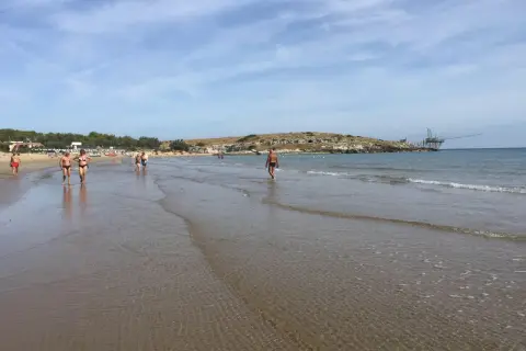 Persone passeggiano sulla battigia di Cala Molinella, con un trabucco sullo sfondo.
