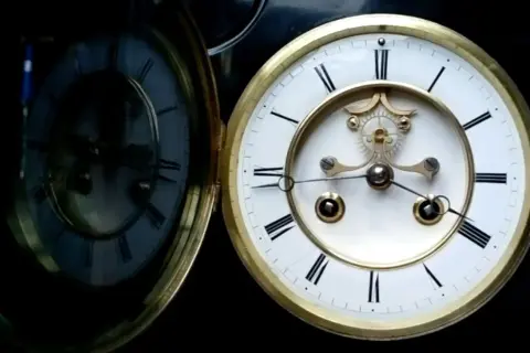Stary zegar wahadłowy spóźnia się. Jego tarcza z rzymskimi cyframi i złote zdobienia odbijają się w lustrzanej powierzchni.