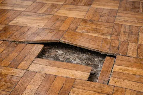 Suelo laminado con tablillas levantadas y un hueco expuesto, evidenciando problemas de humedad o instalación.