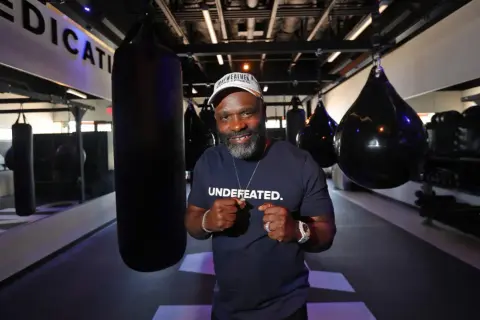 A man in a "UNDEFATED" shirt and Mayweather Boxing + Fitness hat poses with fists up in a gym, ready for Floyd Mayweather training.