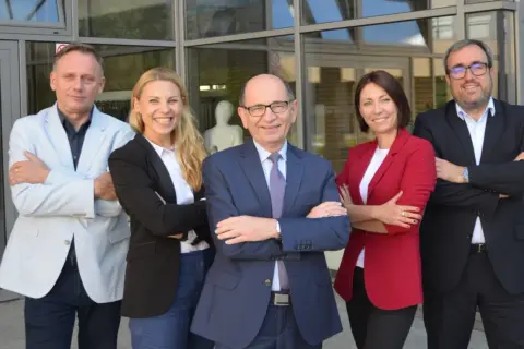 Profesor nadzwyczajny w okularach i jego zespół pozują do zdjęcia przed budynkiem.