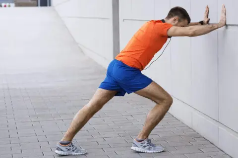 Un homme en tenue de sport fait un étirement mollet contre un mur blanc.