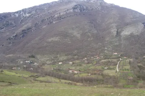 Un paesaggio collinare dove il Cilento mostra case sparse tra alberi spogli e prati verdi, sotto un imponente monte roccioso.