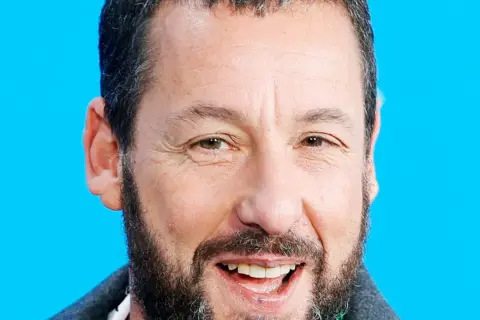 Uśmiechnięty Adam Sandler, gwiazda wielu komediowych filmów, na tle niebieskiego tła.