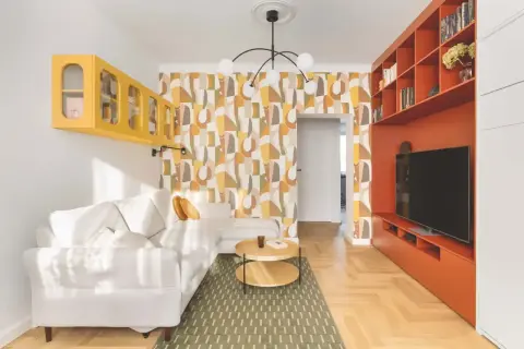 Salon w stylu retro inspiracje: biała kanapa, żółta półka, pomarańczowa zabudowa RTV i geometryczna tapeta tworzą przytulną przestrzeń.