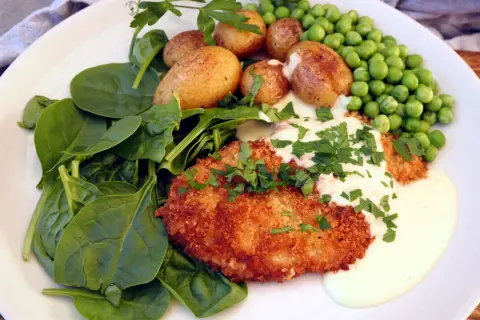 En panerad schnitzel med krämig sås, serverad med färsk spenat, små potatisar och gröna ärtor. En klassisk rätt där såsen till schnitzel lyfter hela smakupplevelsen.