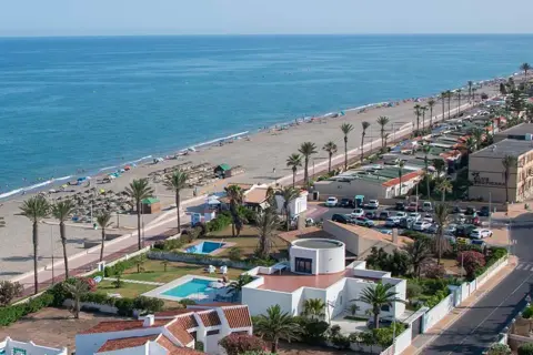 Vista aérea de una playa con sombrillas y gente, palmeras y edificios. Merece la pena Roquetas de Mar.