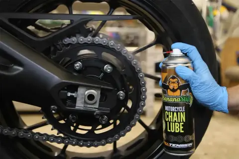 Ręka w niebieskiej rękawiczce aplikuje smar do łańcucha motocyklowego z puszki Banana Slip.