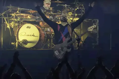 Wokalista Riverside z uniesionymi rękami, basista na scenie, perkusista w tle. Koncertowa atmosfera, publiczność wiwatuje.
