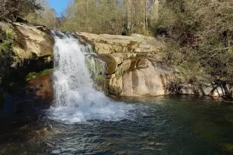 Cascada de agua blanca cayendo sobre rocas, formando pozas de melón en el río. Vegetación frondosa y árboles secos rodean el paisaje.