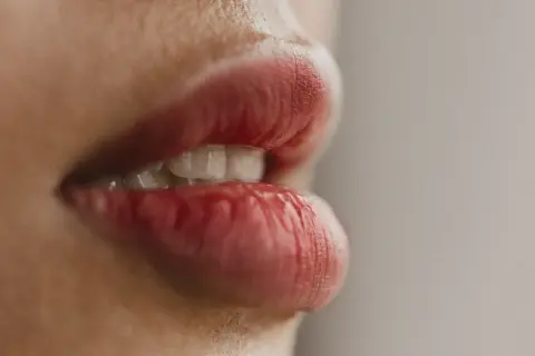 Zbliżenie na usta z widocznymi zębami. Czerwona szminka podkreśla ich kształt, choć widać lekkie suche usta.