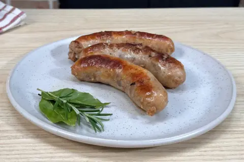 Tre salsicce dorate e succulente, appena cotte in padella, riposano su un piatto con rosmarino e alloro.