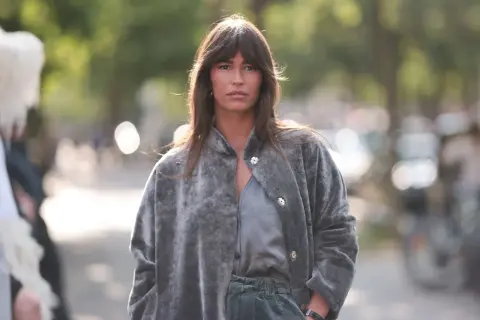 Frau mit curtain bangs und natürlichen Locken trägt einen grauen Kunstpelzmantel und grüne Hose.