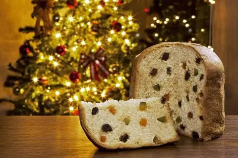 Un panettone, symbole de Noël, avec sa tranche coupée révélant fruits confits et raisins secs. Son origine italienne se célèbre devant un sapin illuminé.