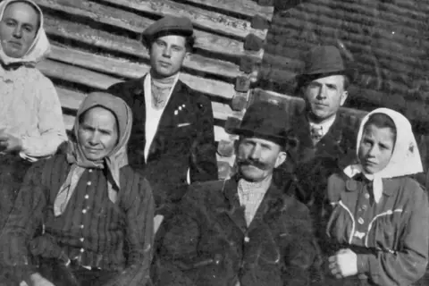 Famille posant devant une maison en bois. Les femmes portent des foulards, les hommes des chapeaux. Un nom de famille slovaque pourrait être associé à cette photo d'ancêtres.