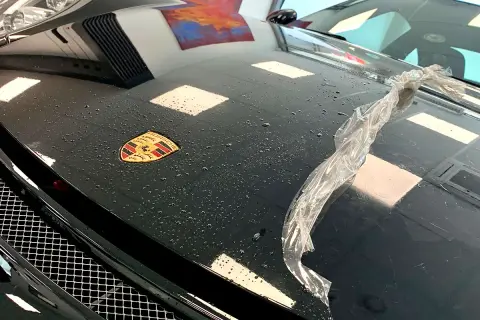 Czarna maska Porsche z logo, na której widać krople wody i fragment folii. Trwa klejenie folii na sucho.