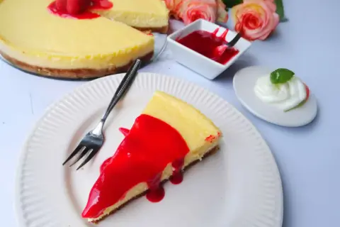 Ein Stück New York Cheesecake mit roter Fruchtsauce und Himbeeren, daneben eine kleine Schüssel Sauce und ein Klecks Sahne.