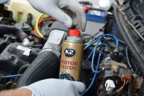 Ręka w rękawiczce trzyma puszkę płukanki do silnika K2 Motor Flush na tle silnika samochodowego.
