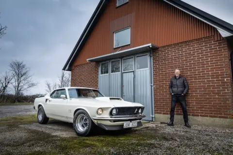 En man står stolt bredvid sin vita Ford Mustang Boss 429 framför en röd tegelbyggnad.