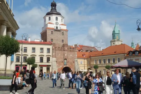 Ludzie spacerują po brukowanym placu litewskim, podziwiając zabytkową wieżę z zegarem i zieloną iglicę w tle.