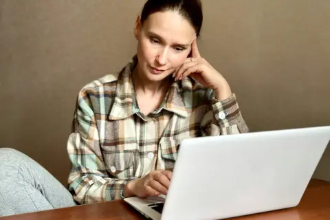 Kobieta w kratę siedzi przy laptopie, zamyślona, z ręką na skroni.
