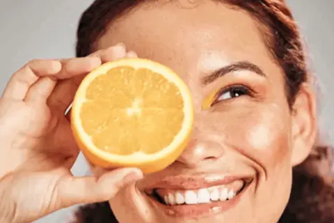 Strahlendes Lächeln mit einer Orangenscheibe vor dem Auge, die Frische und Vitamin C für die Hautpflege symbolisiert.