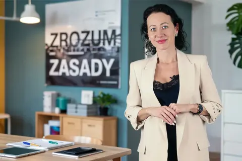 Kobieta w beżowej marynarce, obok niej na biurku leży tablet i notatnik, który może służyć jako księga przychodów i rozchodów.