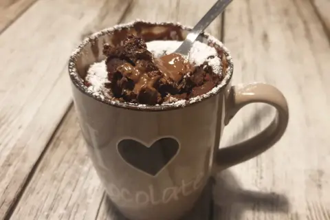 Un delizioso mug cake al cioccolato, appena sfornato, con cuore e scritta "chocolate" sulla tazza, spolverato di zucchero a velo.