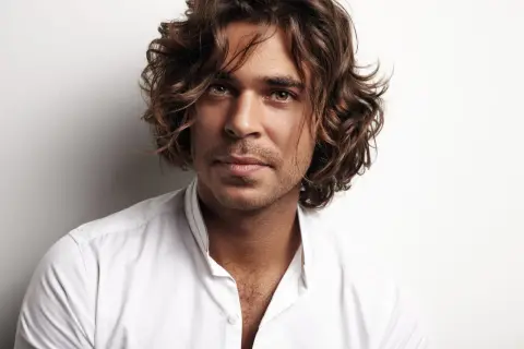 Hombre con peinados media melena, cabello castaño ondulado, mirada intensa y camisa blanca desabrochada.