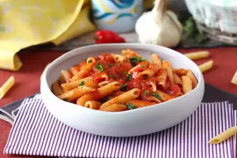 En skål med pasta arrabiata, toppad med färsk persilja. En vitlöksklyfta och en röd chili syns i bakgrunden.