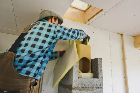 Pracownik w kratkę montuje elementy do budowy komina, używając wełny mineralnej.