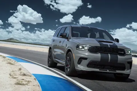 Szary Dodge Durango SRT Hellcat z czarnymi pasami pędzi po torze wyścigowym pod błękitnym niebem z chmurami.